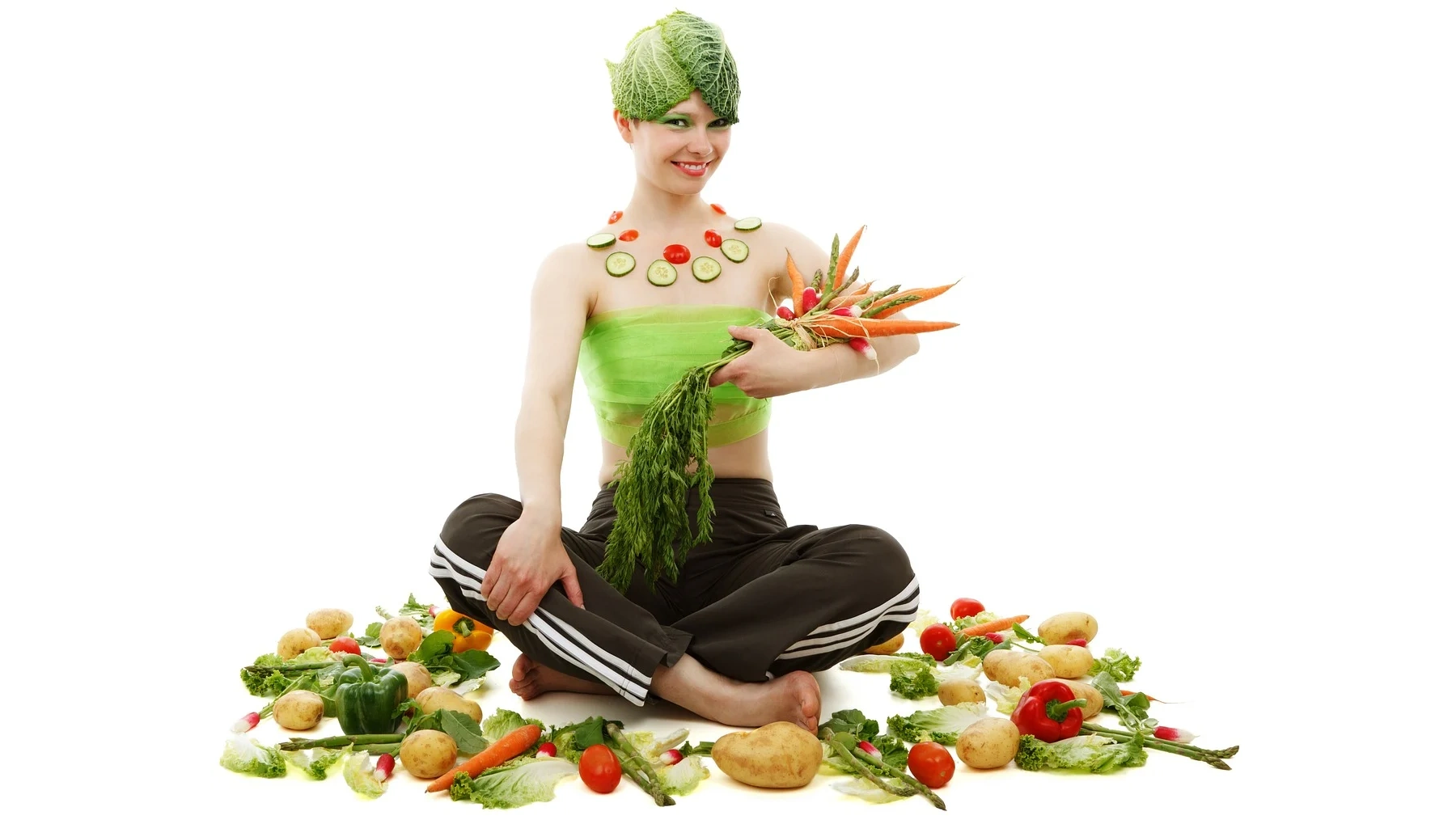 Young woman sits surrounded by vegetables, holds them in her arms and has cabbage leaves on her head.